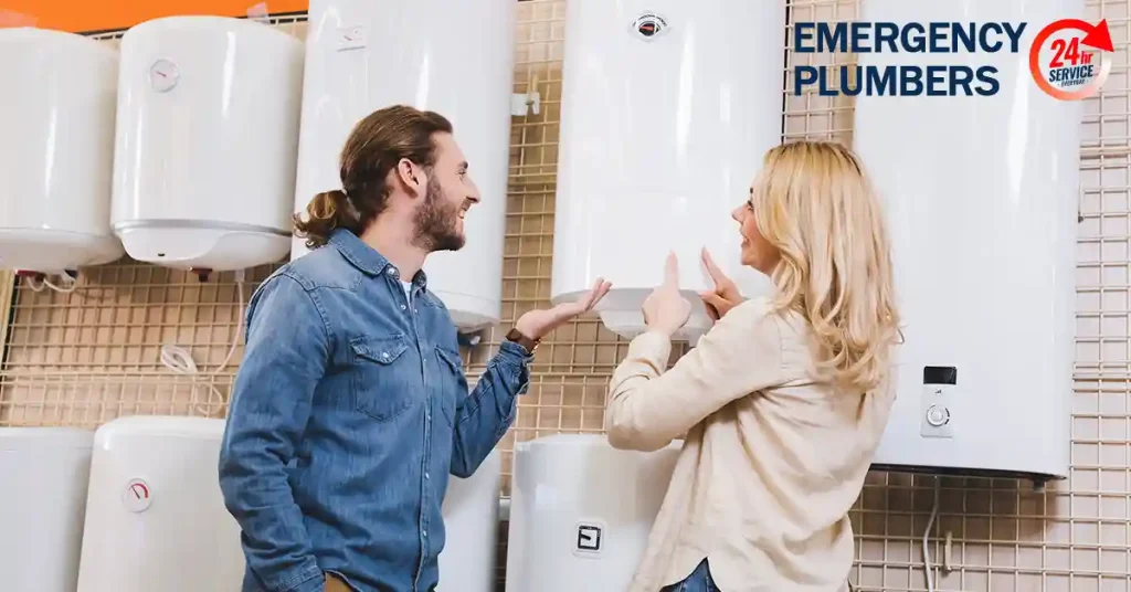 A man and woman discuss options while standing in front of wall-mounted water heaters in a plumbing store with a sign reading "Emergency Plumbers.