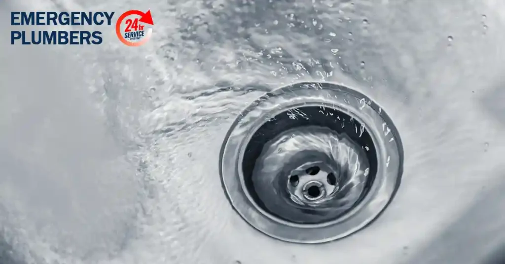 Image of a sink drain with water swirling down, accompanied by the text "EMERGENCY PLUMBERS" and a "24h Service" logo in the top left corner.