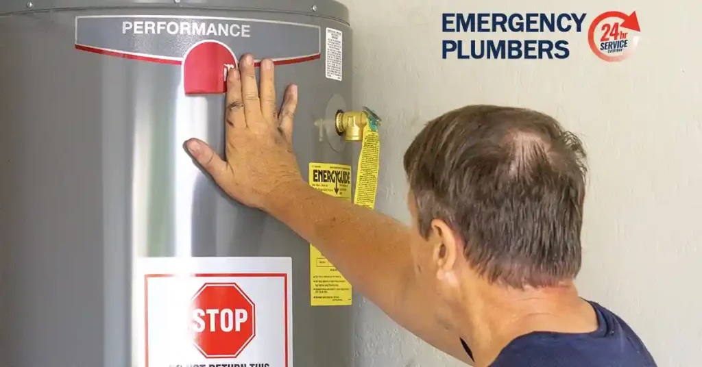 A person adjusts a water heater next to a wall with a sign that reads "EMERGENCY PLUMBERS 24/7 SERVICE.