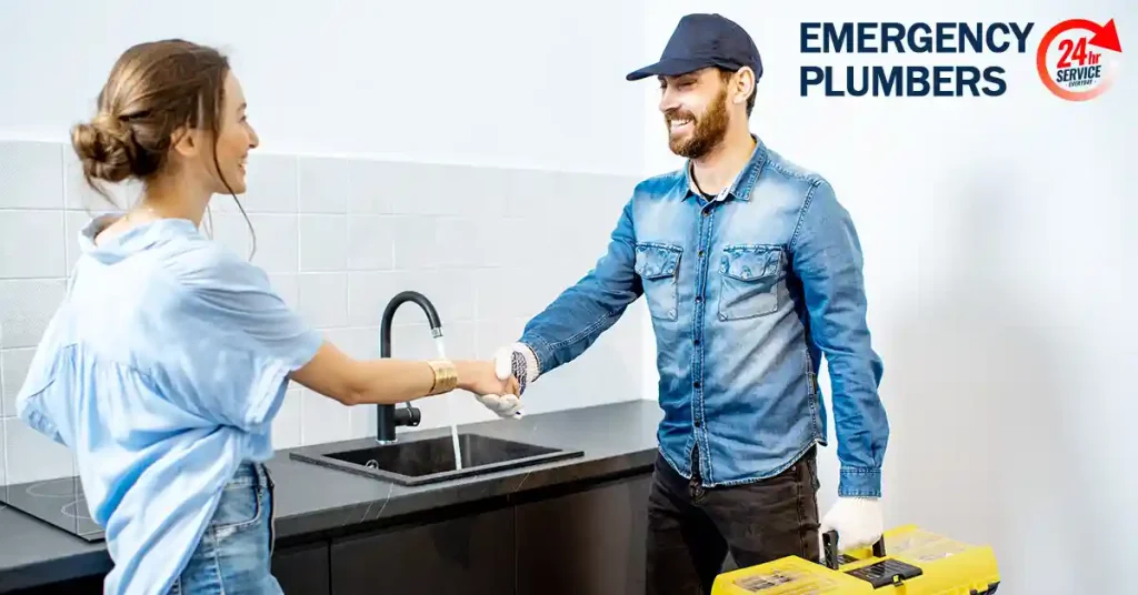 A plumber wearing a cap shakes hands with a woman standing by a kitchen sink. A tool case is in the plumber's other hand. Visible text reads "Emergency Plumbers" and "24/7 Service.