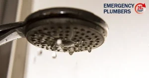 Close-up of a showerhead with water droplets falling. "EMERGENCY PLUMBERS" and "24/7 SERVICE" text with a red logo are visible in the background.