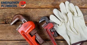 Two pipe wrenches and a pair of work gloves on a wooden surface. Text on the image reads "Emergency Plumbers" with a small circular emblem in the top left corner.