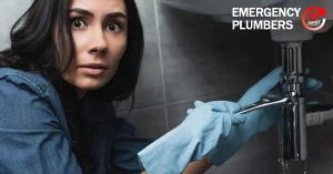Person wearing blue gloves holding a wrench under a sink with a concerned expression. The text "EMERGENCY PLUMBERS" and a service logo are visible in the upper right corner.
