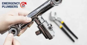 A person holding a plumbing valve with tools in the background. The text reads "Emergency Plumbers 24-hour Service.