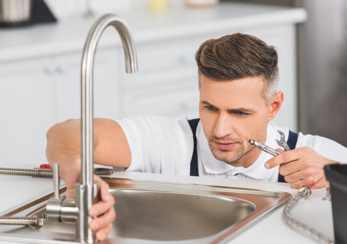 A man in blue overalls, delivering plumbing services, is repairing a sink in a kitchen. He is holding a wrench in one hand and adjusting the faucet with the other.