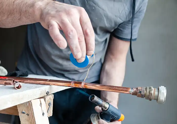 A plumber is repairing a burst copper pipe with solder wire and a blowtorch on a wooden workbench.