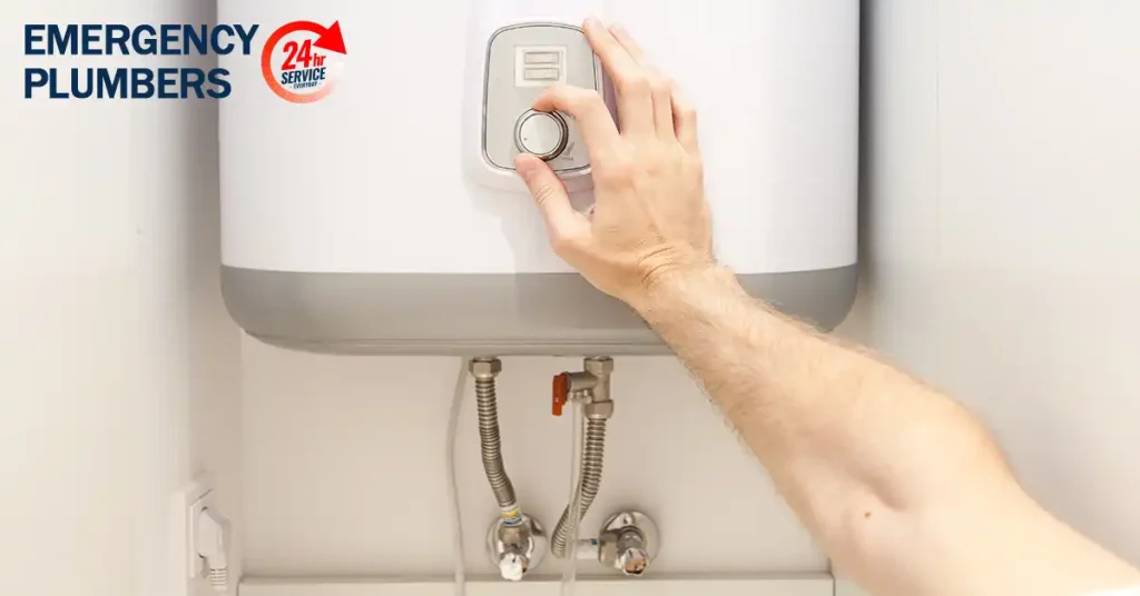 Hand adjusting the control dial on a wall-mounted water heater, with "Emergency Plumbers 24hr Service" logo in the top left corner.